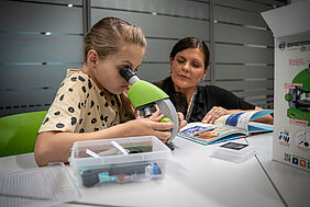 Ein Mädchen mikroskopiert während ie Mutter daneben hockt und zusieht