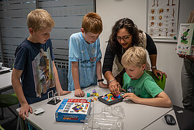 Ein Junge spielt ein Spiel währedessen andere Kinder und eine Frau um den Tisch herum stehen